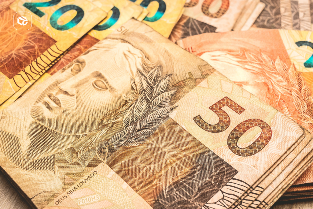 Close-up de várias cédulas de real brasileiro sobrepostas, com destaque para uma nota de R$ 50 em primeiro plano. A imagem evidencia detalhes da impressão da cédula, incluindo a efígie da República, elementos de segurança e a numeração do valor. Ao fundo, aparecem parcialmente notas de R$ 20 e outras denominações. No canto superior esquerdo há um pequeno logotipo branco dentro de um quadrado com cantos arredondados.