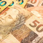 Close-up de várias cédulas de real brasileiro sobrepostas, com destaque para uma nota de R$ 50 em primeiro plano. A imagem evidencia detalhes da impressão da cédula, incluindo a efígie da República, elementos de segurança e a numeração do valor. Ao fundo, aparecem parcialmente notas de R$ 20 e outras denominações. No canto superior esquerdo há um pequeno logotipo branco dentro de um quadrado com cantos arredondados.